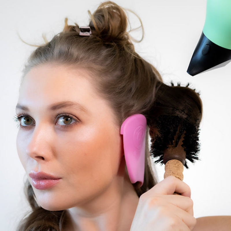Full frontal photo of a white woman using a blow dryer to dry her hair while wearing pink color Miss Careful brand of Ear Covers on her ears