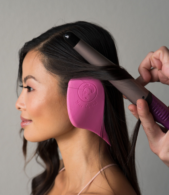 woman with long dark hair wearing Miss Careful pink color ear covers while curling hair